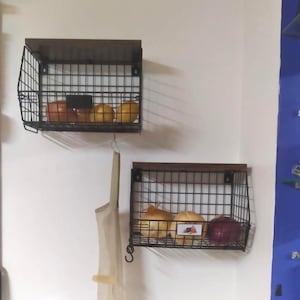 May include: Two wall-mounted wire baskets with wooden tops. The baskets are filled with various fruits and vegetables. The baskets are black and the wooden tops are brown. A beige bag hangs below one of the baskets.