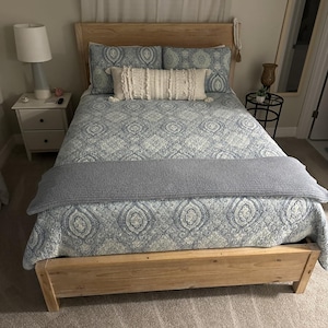 May include: A light wood bed frame with a patterned blue and white quilt and matching pillows. A gray knitted throw is draped across the foot of the bed. A white nightstand with a lamp is on the left.