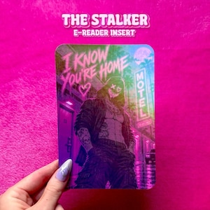 May include: A rectangular e-reader insert featuring a neon-toned illustration of a hooded figure with a glowing mask. The text reads I KNOW YOU'RE HOME above the figure and MOTEL on a vertical sign. The color palette includes vibrant pinks, purples, and greens.
