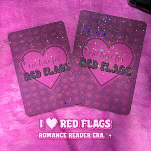 Può includere: Due carte rosa con un design a cuore e la scritta "I'm here for RED FLAGS". Le carte hanno un effetto glitterato e la frase "I ❤️ RED FLAGS ROMANCE READER ERA" è in basso.