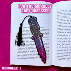 Pode incluir: Um marcador de página em forma de adaga com um degradê preto e roxo, gotas rosas, detalhes de estrelas e o texto "Morally Grey Is My Type" no centro. O marcador inclui uma borla preta presa ao cabo e está sobre um livro aberto.