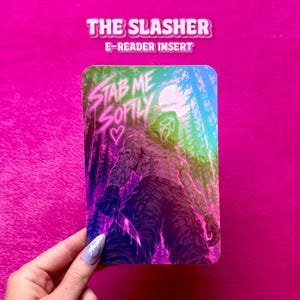 Peut inclure: Un insert pour liseuse rectangulaire présentant une illustration holographique multicolore d'une silhouette masquée dans une forêt. La silhouette porte un masque à capuche et tient un couteau. Le texte "STAB ME SOFTLY" et un cœur sont imprimés en rose néon.