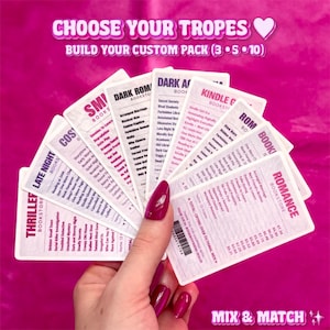 Op de afbeelding: Een hand houdt een waaier van witte kaarten met verschillende boekgenres en titels, waaronder "Romance Bookstore" en "Thriller Bookstore". De kaarten bevatten tekst als "Choose Your Tropes" en "Mix & Match" op een roze achtergrond.