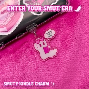 Peut inclure: Un charm en acrylique transparent avec un motif de fantôme tenant un livre, avec des yeux en forme de cœur et une bulle de pensée disant "IS THAT SMUT AGAIN ?". Le charm est attaché à une coque de téléphone. Le texte sur la coque dit "ENTER YOUR SMUT ERA" et "SMUTY KINDLE CHARM".