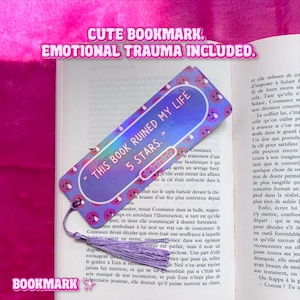 Peut inclure: Un marque-page rectangulaire violet orné d'un motif répétitif d'emojis en pleurs. Le texte central indique "THIS BOOK RUINED MY LIFE 5 STARS." avec un graphique de cinq étoiles. Un pompon violet clair est attaché au coin inférieur.