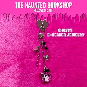 Peut inclure: Une breloque pour liseuse argentée avec un personnage fantôme portant un chapeau rose et des lunettes de soleil, ornée de perles noires et roses, d'une petite citrouille et d'une chaîne. Le texte indique THE HAUNTED BOOKSHOP HALLOWEEN 2K26 GHOSTY E-READER JEWELRY.