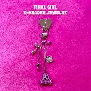 Peut inclure: Un accessoire à breloques en métal argenté avec un sommet en forme de cœur, une breloque téléphone, une breloque télévision et un pendentif masque de fantôme avec le texte "WHAT'S YOUR FAVORITE SCARY BOOK?" orné de perles roses et noires.