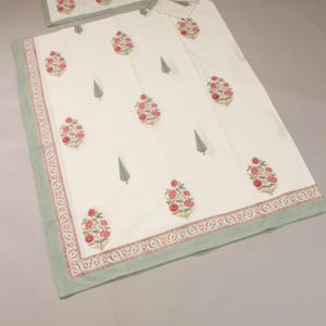 Könnte beinhalten: Weißes Bettwäsche-Set mit einem Blumenmuster in Rot und Rosa, akzentuiert mit grünen Blättern. Das Set beinhaltet eine Decke, einen Kissenbezug und ein kleineres rechteckiges Stück, alles mit einem grünen Rand.