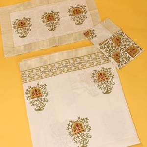 Könnte beinhalten: Ein Set aus weißer Tischwäsche mit einem sich wiederholenden olivgrünen Blumenmuster und kleinen roten Akzenten. Die Kollektion umfasst ein rechteckiges Platzset, eine gefaltete Serviette und ein größeres rechteckiges Tuch mit gestreiften Rändern.