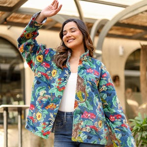 Könnte beinhalten: Blau gesteppte Jacke mit einem lebendigen Blumen- und Affenmuster. Die Jacke hat einen Stehkragen und wird offen über einem weißen Oberteil und einer blauen Jeans getragen. Der Gesamtstil ist lässig und farbenfroh.