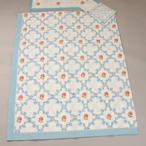 Op de afbeelding: Een witte beddengoedset met een blauwe rand en een herhalend patroon van blauwe vierkanten en roze rozen. De set bevat een groot deken, een rechthoekige kussensloop en een kleinere vierkante kussensloop.
