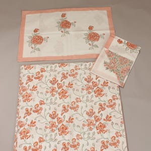 May include: A white floral print tablecloth, placemat, and napkin set. The tablecloth features a repeating pattern of orange flowers and green stems. The placemat and napkin have a similar floral design with a pink border. The set is made of a soft, lightweight fabric.