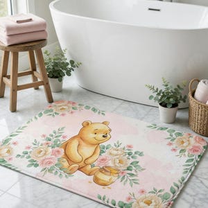 May include: A rectangular bath mat featuring a cartoon bear with a honey pot, surrounded by floral elements. The mat has a pink watercolor background and a white border. The mat is designed for bathroom use.