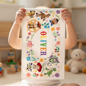 May include: White rectangular towel featuring colorful illustrations of characters and flowers. The name "OLIVER" is vertically printed in rainbow colors. The towel has a border of flowers and characters from a popular animated movie.