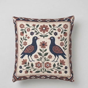 May include: Square decorative pillow with a cream-colored background featuring two stylized blue birds facing each other. The birds are surrounded by a floral design in shades of brown, green, and cream. The pillow has a patterned border.