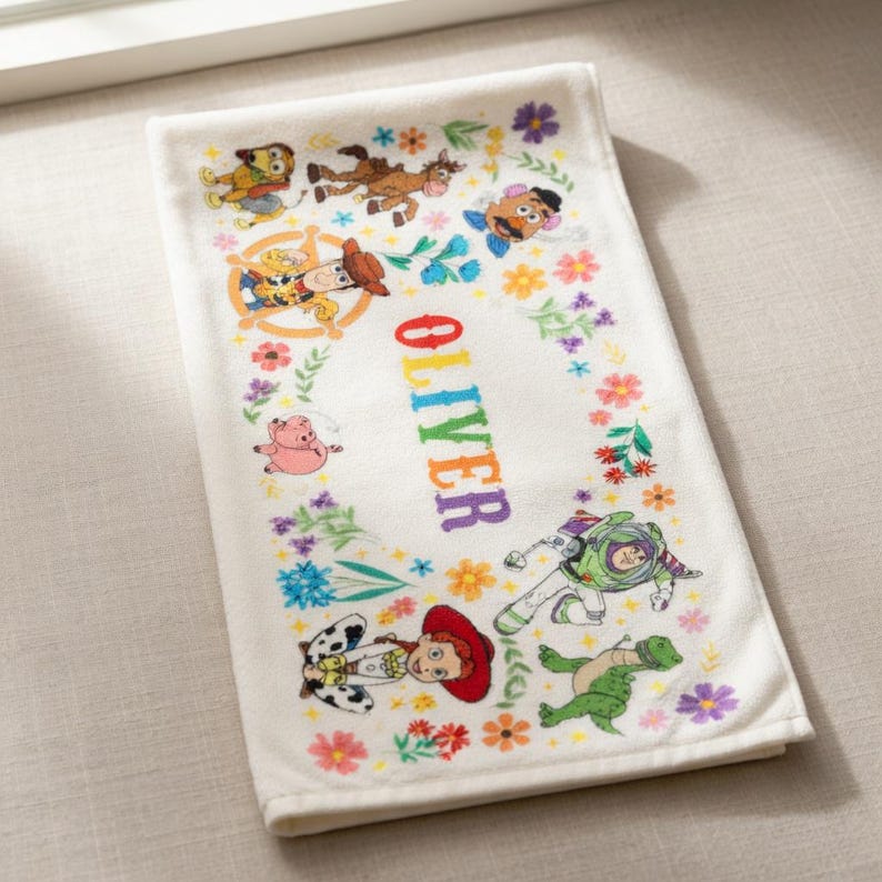 May include: White rectangular towel featuring colorful illustrations of Toy Story characters and the name "OLIVER" in rainbow letters. The towel is decorated with floral accents.