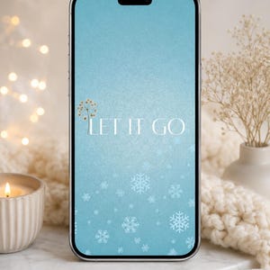 May include: A smartphone displays a light blue background with snowflakes and the text "LET IT GO". The phone is on a white marble surface, with a lit candle and decorative flowers in the background.