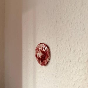 May include: A small, oval ceramic wall art piece. The art has a glossy finish with a marbled pattern of deep red and white. The piece is mounted on a textured white wall.