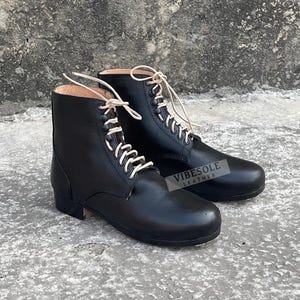 May include: A pair of black leather ankle boots with white laces. The boots have a classic design with a rounded toe and a low heel. The boots are displayed on a concrete surface. The text "VIBESOLE LEATHER" is visible on the side of one boot.