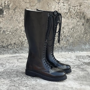 May include: A pair of black knee-high leather boots featuring a full-length lace-up front with black eyelets and black laces. The boots have a rounded toe, a thick lugged rubber sole, and a small logo patch reading VIBESOLE LEATHER on the side.
