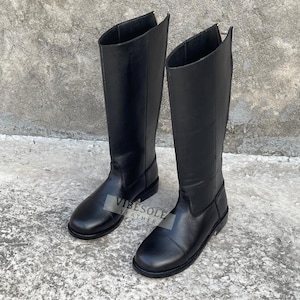 May include: A pair of black knee-high leather boots with a rounded toe, a low heel, and a vertical zip closure along the back. The text VIBESOLE LEATHER is printed in white on the side of the left boot. The boots feature a smooth finish.