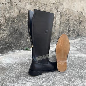 以下が含まれることがあります： 黒い革製のニーハイブーツ。サイドジッパーとゴールドカラーのジッパープルが付いています。ブーツは、薄茶色のソールの隣に表示されています。「Vibesole Leather」というブランド名がブーツの側面に表示されています。