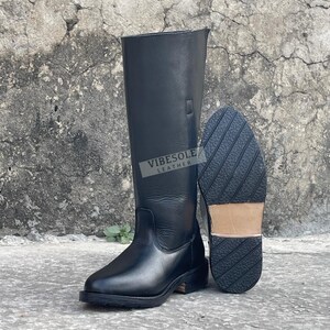 May include: Black leather riding boots with a tall shaft and a textured sole. One boot is upright, and the other is lying on its side, displaying the sole. The boots have the text "VIBESOLE LEATHER" on the side.