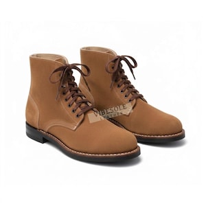 May include: A pair of tan suede ankle boots featuring dark brown laces, metal eyelets, and a black sole with visible white stitching. The side of the boot displays a small rectangular tag with the text VIBESOLE LEATHER printed in black.