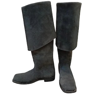 Pode incluir: Um par de botas de camurça cinza escuro com punho dobrado. As botas têm bico quadrado e salto baixo. O material de camurça confere às botas um aspecto macio e texturizado. Estas botas são adequadas para fantasias ou reconstituições históricas.