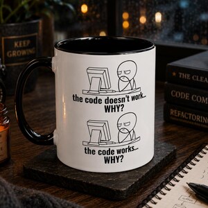 Op de afbeelding: Een witte keramische mok met een zwarte binnenkant en handvat, voorzien van een zwarte lijntekening van een persoon achter een computer. De tekst luidt: the code doesn't work... WHY? en the code works... WHY? De mok is cilindervormig.
