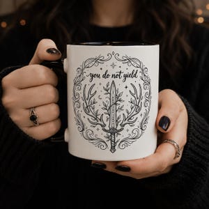 May include: White ceramic mug with a black interior and handle. The mug features a black and white illustration of a sword, antlers, and the text "you do not yield". The design is surrounded by a decorative border.