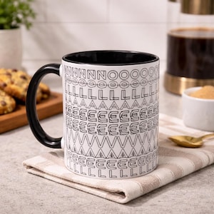 Peut inclure: Tasse en céramique blanche avec une anse et un bord noirs, ornée d'un motif répété de lettres et de symboles noirs. La tasse est posée sur une serviette à rayures beiges et blanches. Des cookies aux pépites de chocolat sont visibles en arrière-plan.