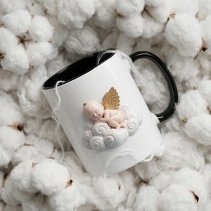 May include: White ceramic mug with a black interior and handle. The mug features a sleeping baby angel on a cloud with gold wings. The mug is surrounded by white cotton bolls.