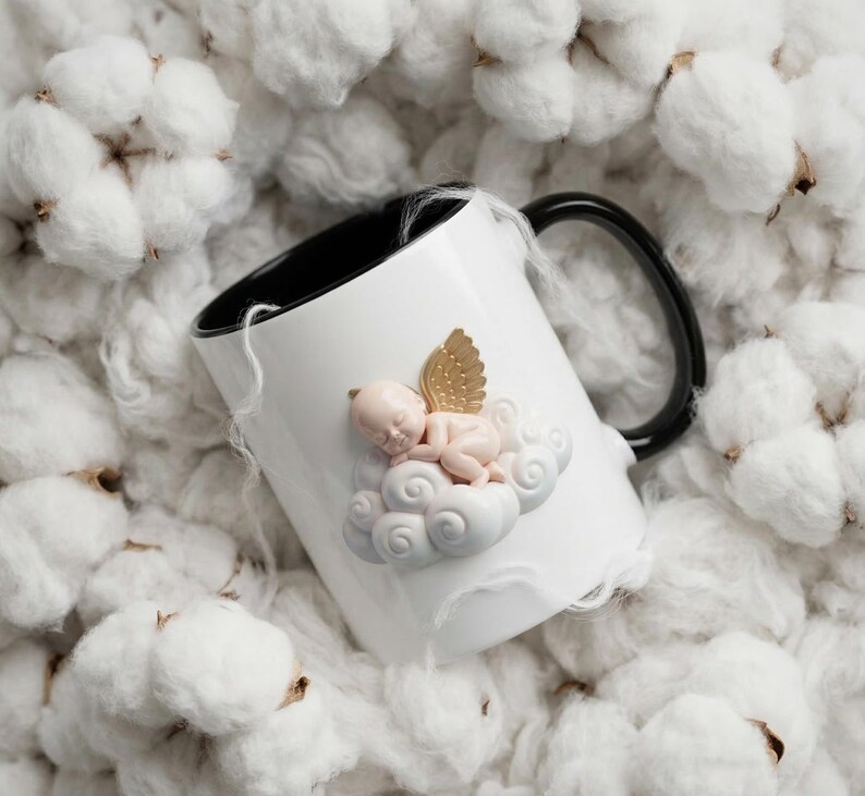 May include: White ceramic mug with a black interior and handle. The mug features a sleeping baby angel on a cloud with gold wings. The mug is surrounded by white cotton bolls.