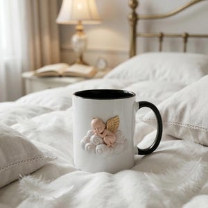 May include: White ceramic mug with a black interior and handle. The mug features a 3D relief of a sleeping cherub with gold wings on a cloud. The mug is resting on a white bed with pillows.