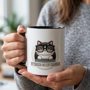 Pode incluir: Caneca de cerâmica branca com interior e alça pretos. A caneca apresenta um gato de desenho animado usando óculos e digitando em um teclado. O texto "IT WORKS ON MY MACHINE" está abaixo da ilustração.