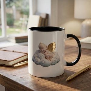 May include: A white ceramic mug with a black interior and handle. The mug features a sleeping cherub on a cloud design with gold wings. The mug is on a wooden surface.