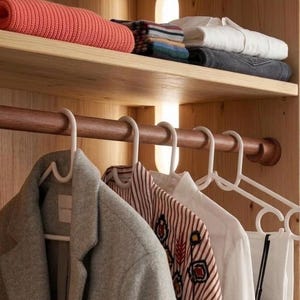 May include: A wooden closet with clothes hanging on hangers. A gray coat, a patterned blouse, and a white shirt are visible. Shelves above hold folded clothes, a rolled blanket, and other items.