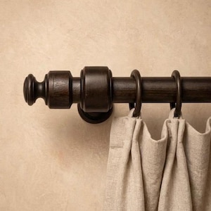 May include: A dark brown curtain rod with decorative finials and rings, holding up a beige linen curtain. The rod is mounted on a light beige wall, creating a neutral and elegant aesthetic.