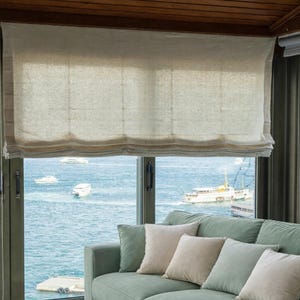 May include: A light beige linen Roman blind is partially lowered over a window overlooking a body of water with boats. A light green sofa with beige and light green cushions is in front of the window.