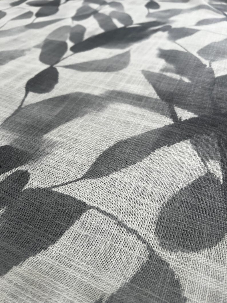 May include: Close-up of a light-colored fabric with a repeating pattern of gray leaves. The leaves have a watercolor effect, with varying shades of gray. The fabric has a subtle, textured appearance.