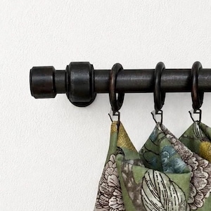 May include: Dark brown metal curtain rod with rings, holding green floral patterned curtains. The rod has a decorative end piece and is mounted on a white wall. The curtains have a mix of green, white, and brown floral designs.