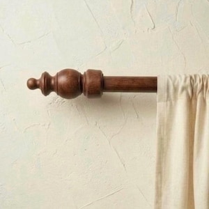 May include: A dark brown wooden curtain rod with decorative finials and a section of a cream-colored curtain. The rod is mounted against a textured, off-white wall, creating a classic and elegant look for window treatments.