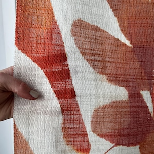 May include: Close-up of a textured fabric with a pattern of large, abstract shapes in shades of orange and rust against a white background. The fabric has a woven appearance, and the design is reminiscent of leaves or organic forms.