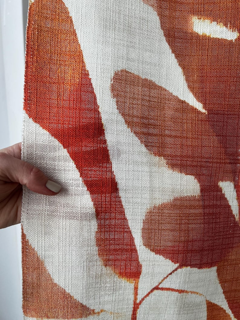 May include: Close-up of a textured fabric with a pattern of large, abstract shapes in shades of orange and rust against a white background. The fabric has a woven appearance, and the design is reminiscent of leaves or organic forms.