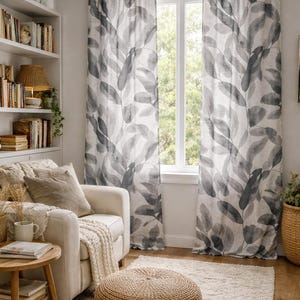May include: A living room scene featuring leaf-patterned curtains in shades of gray and white. A white armchair with a beige throw and a round, woven ottoman sit on a cream-colored rug. A wooden side table holds books and a mug.