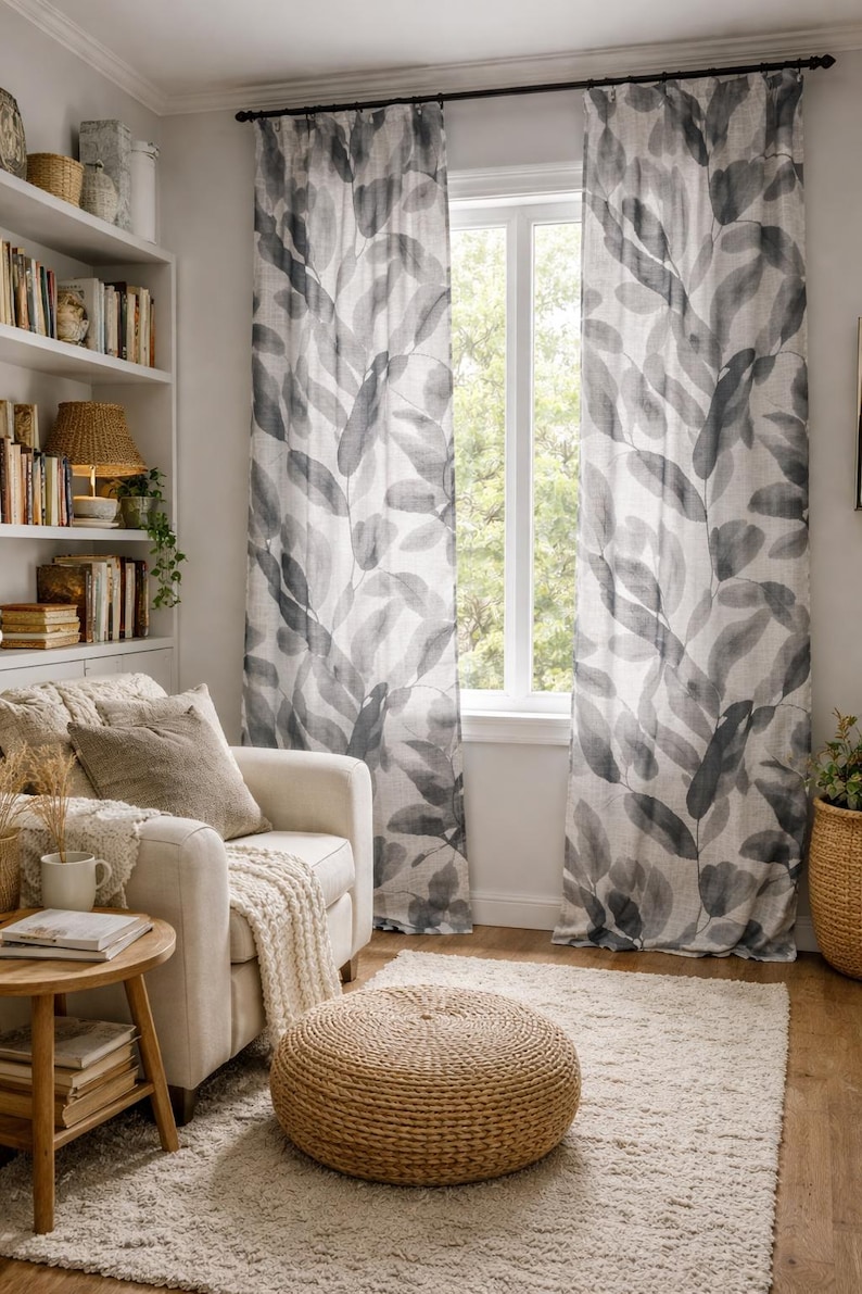 May include: A living room scene featuring leaf-patterned curtains in shades of gray and white. A white armchair with a beige throw and a round, woven ottoman sit on a cream-colored rug. A wooden side table holds books and a mug.