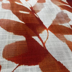 May include: Close-up of a white fabric with a repeating pattern of large, abstract leaves in shades of orange and rust. The leaves have a watercolor-like effect, with soft edges and varying levels of opacity. The fabric has a subtle, textured weave.