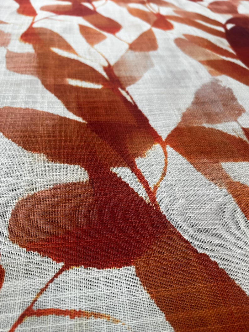 May include: Close-up of a white fabric with a repeating pattern of large, abstract leaves in shades of orange and rust. The leaves have a watercolor-like effect, with soft edges and varying levels of opacity. The fabric has a subtle, textured weave.