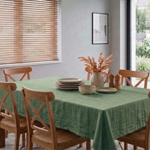 Könnte beinhalten: Ein Esstisch mit einer grünen Tischdecke und Holzstühlen. Auf dem Tisch stehen Teller, eine Schüssel und eine Vase mit Trockenblumen. Im Hintergrund ist ein Fenster mit Jalousien zu sehen. Der Raum hat eine neutrale Farbgebung.